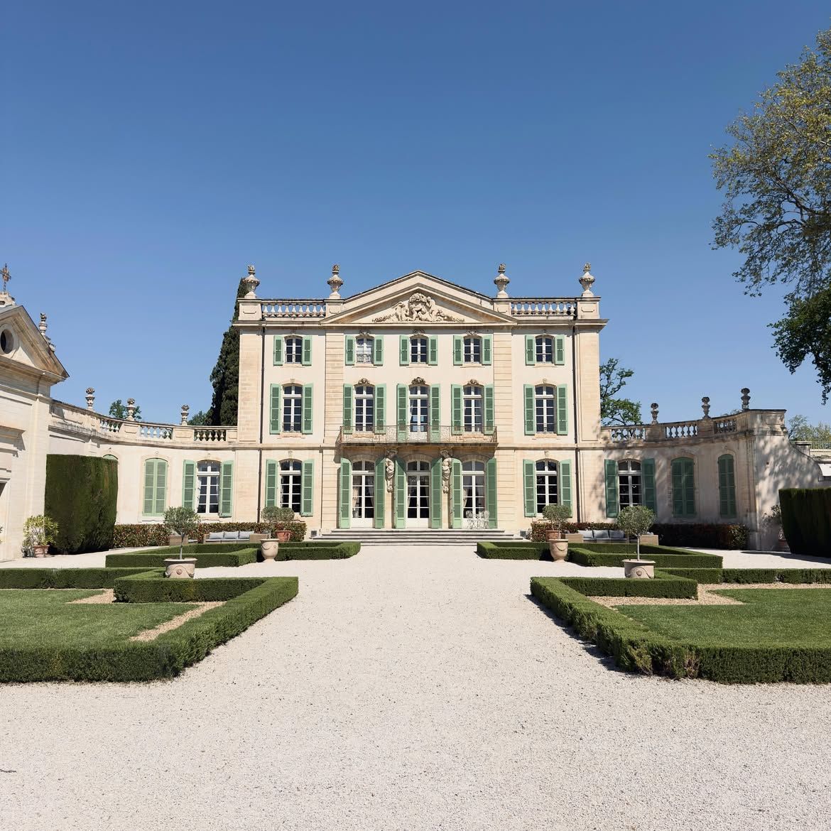 Grand stone mansion with green shutters, formal gardens, and a gravel courtyard under a clear blue sky