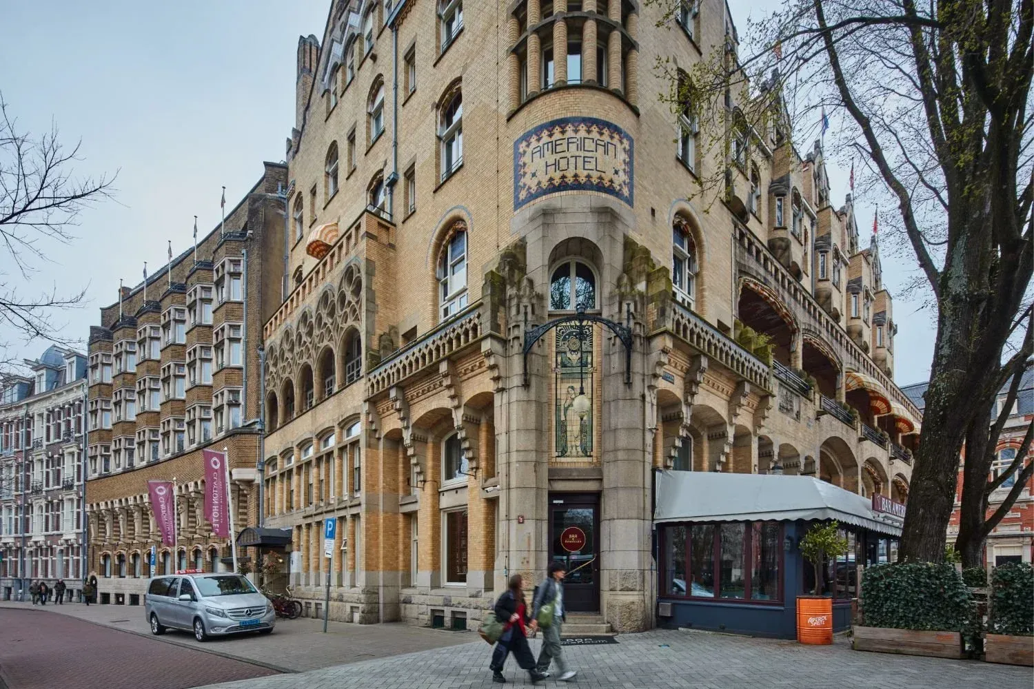 Corner building with ornate facade on a city street, cars parked nearby, and pedestrians under bare trees.