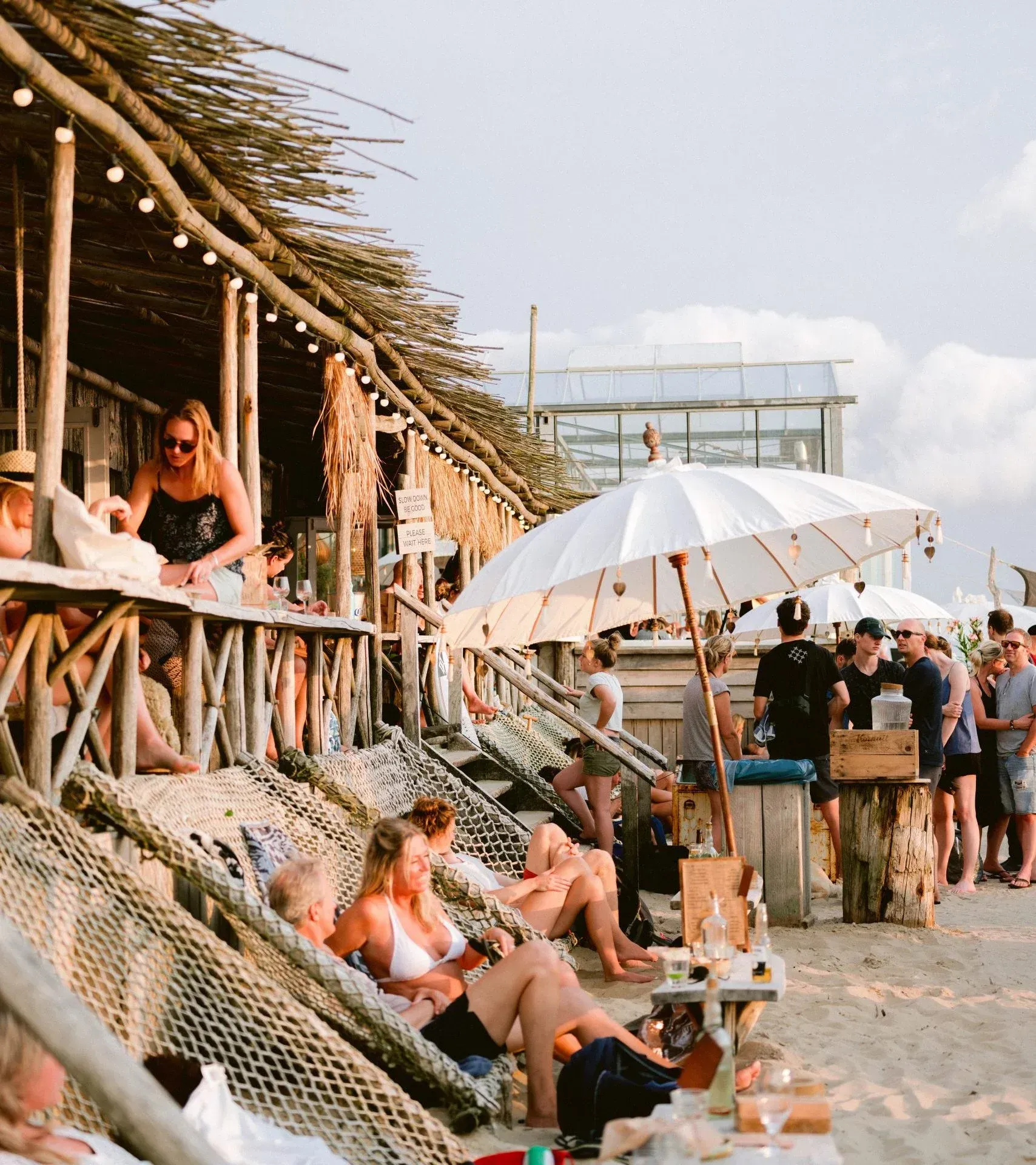 Beachside bar with people lounging on chairs under a white umbrella near rustic wooden huts