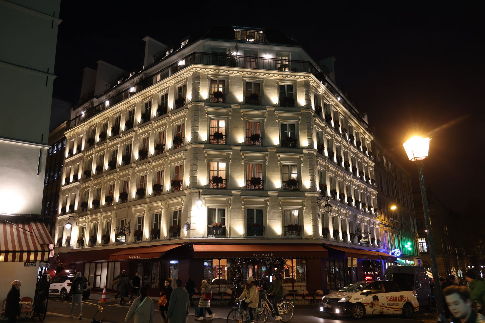 Nighttime view of a brightly lit corner building with balconies and street traffic below