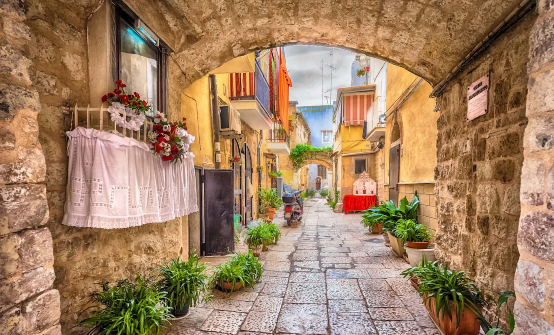 Narrow cobblestone alley under a stone arch, lined with flowers and colorful buildings after rain.
