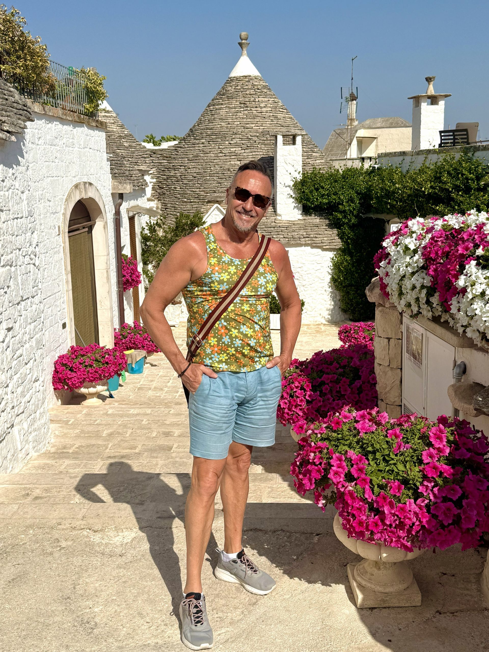Person posing on a sunlit stone path by white walls and pink flowers in a rustic courtyard, smiling in sunglasses.