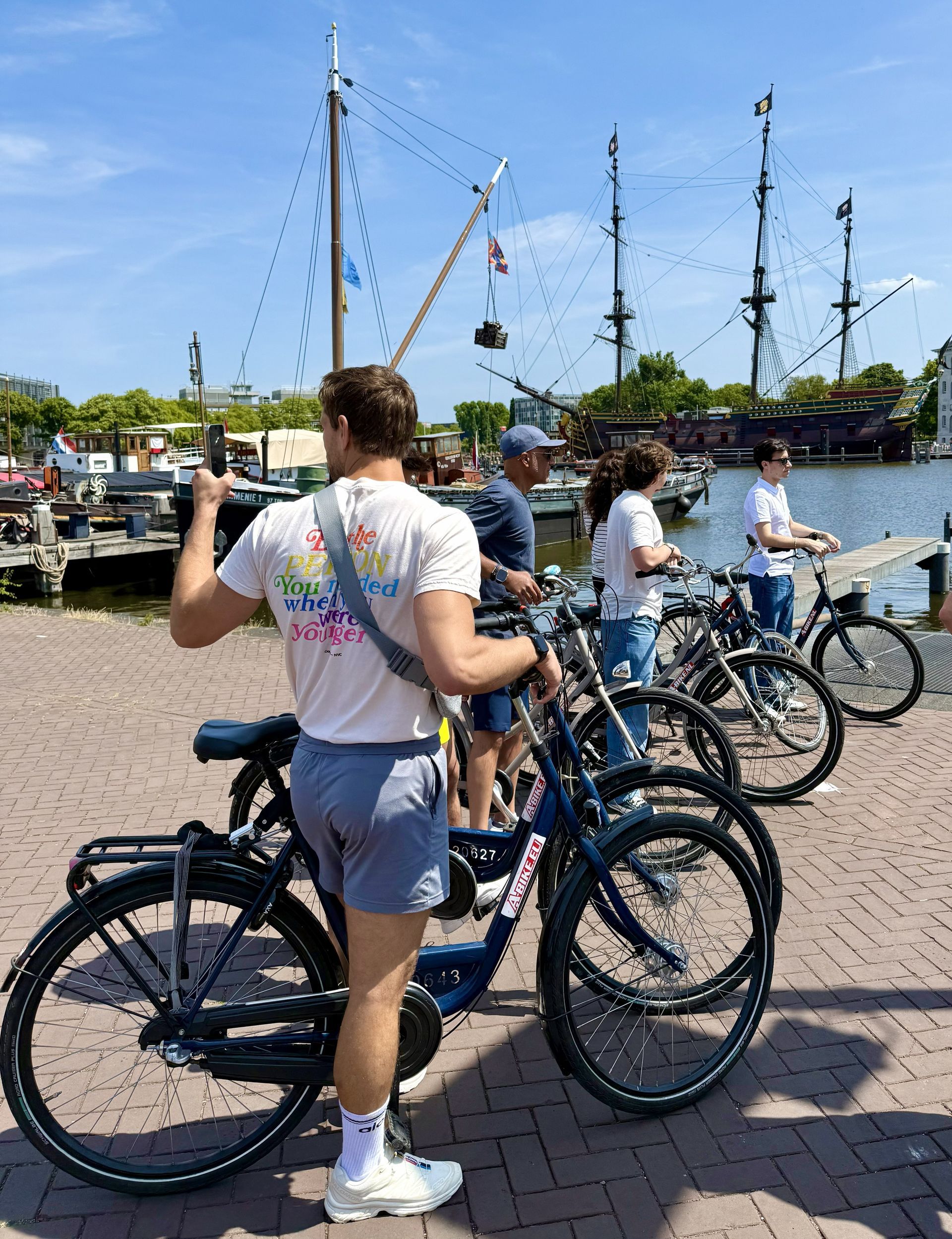 People with bicycles on a sunny harbor dock, with boats and masts in the background.