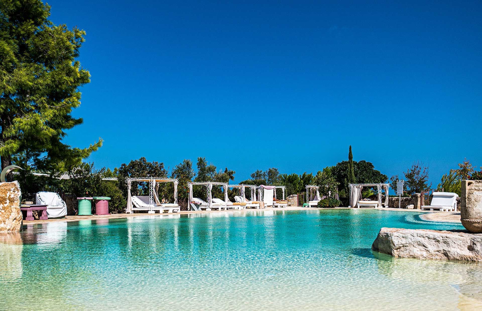 Sunny resort pool with turquoise water, white lounge chairs, and trees under a clear blue sky