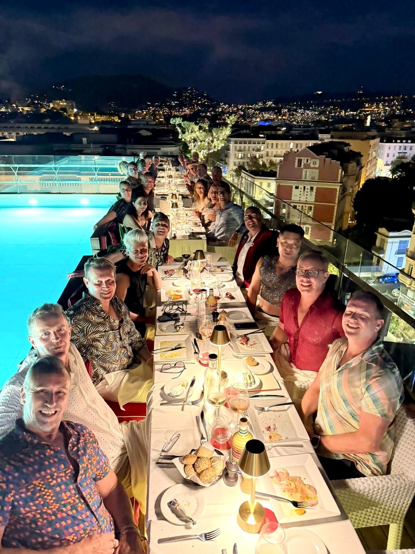 Group dining at a rooftop table at night, with city lights and a pool glowing in the background.