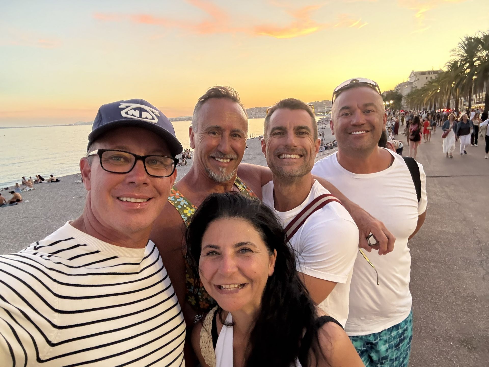 Five smiling friends taking a selfie on a beach at sunset, with the ocean and palm trees behind them.