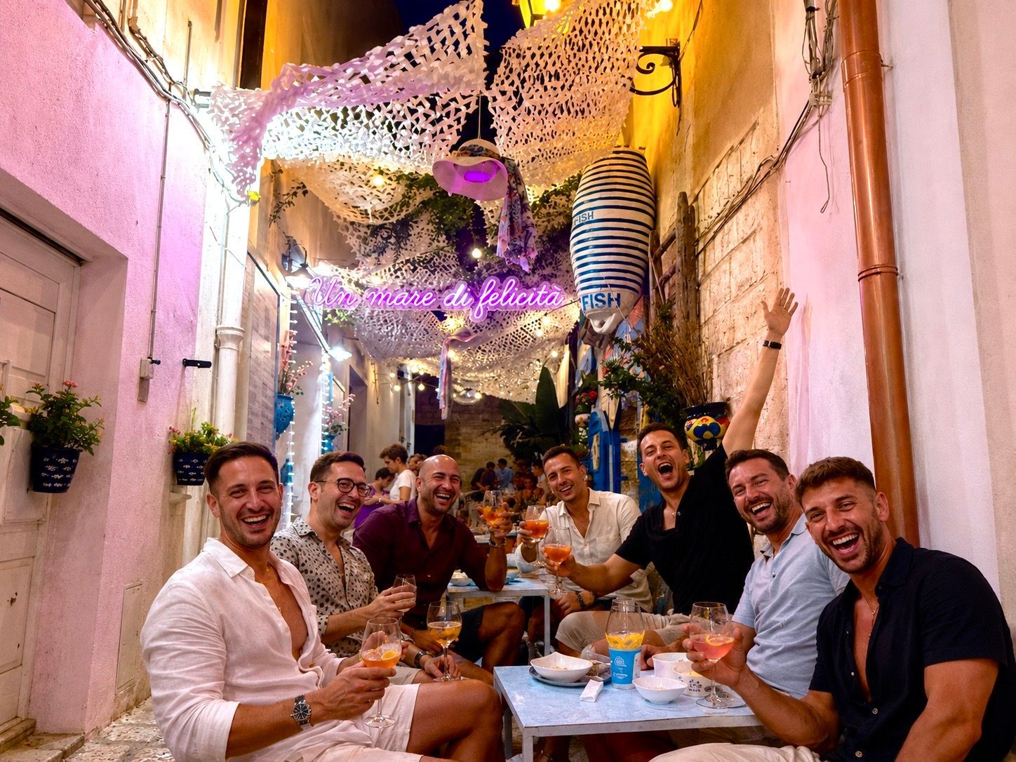 Friends dining at a lively lantern-lit street restaurant, smiling and raising drinks at a shared table