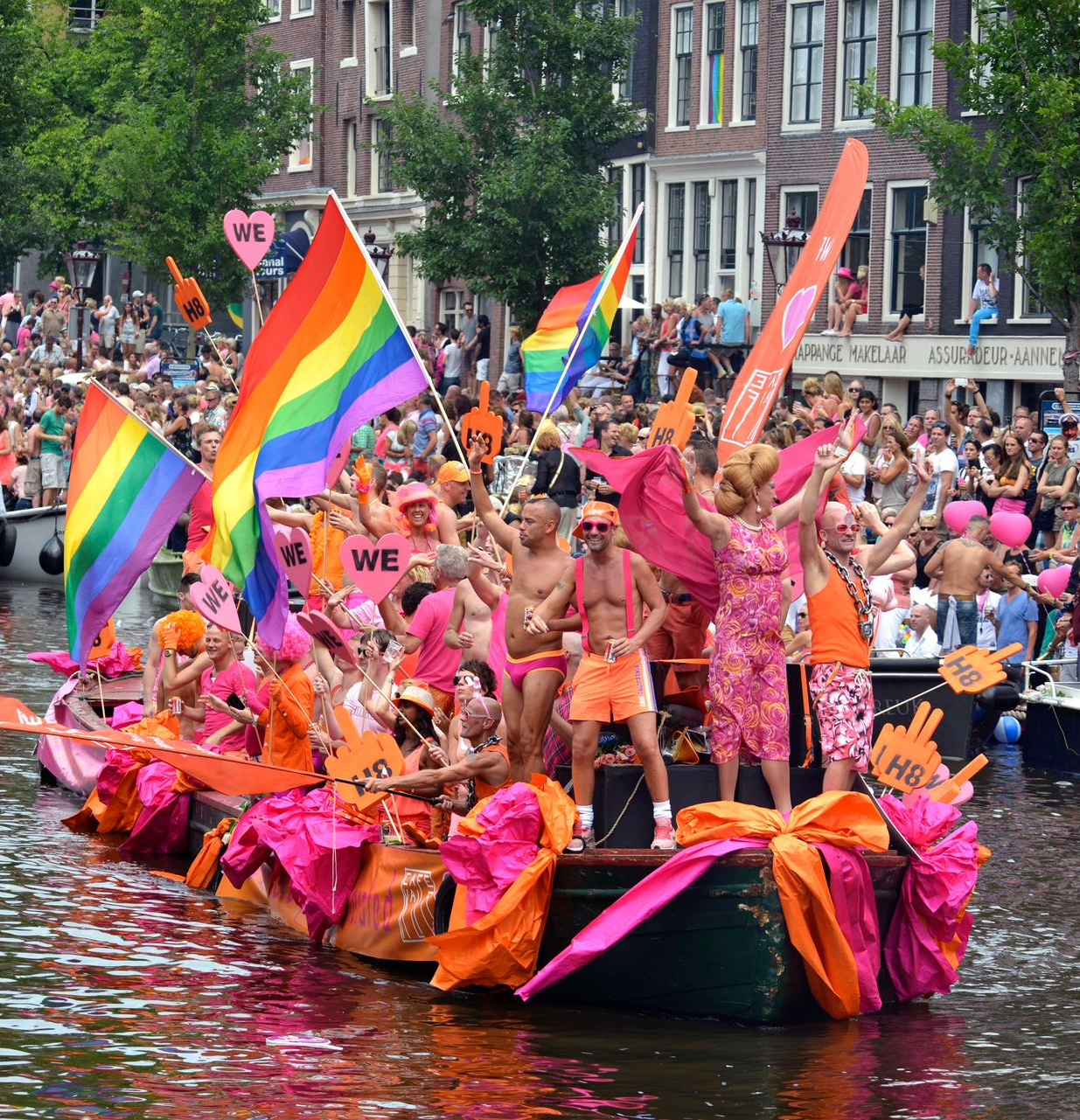 Colorful Pride parade boat on a canal, crowded with people waving rainbow flags and wearing pink costumes.