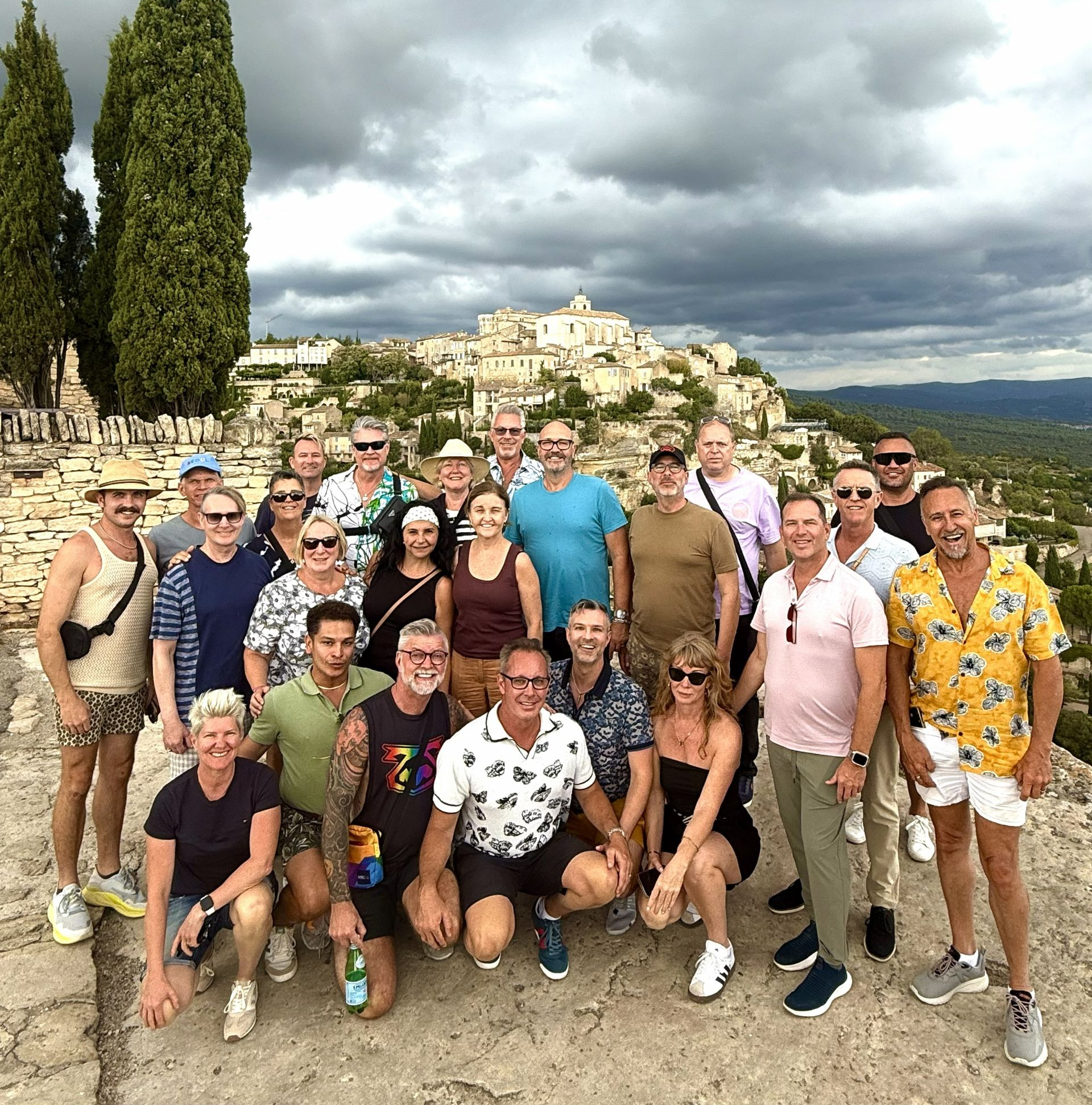 Group of tourists posing outdoors with a hillside village and stormy clouds in the background