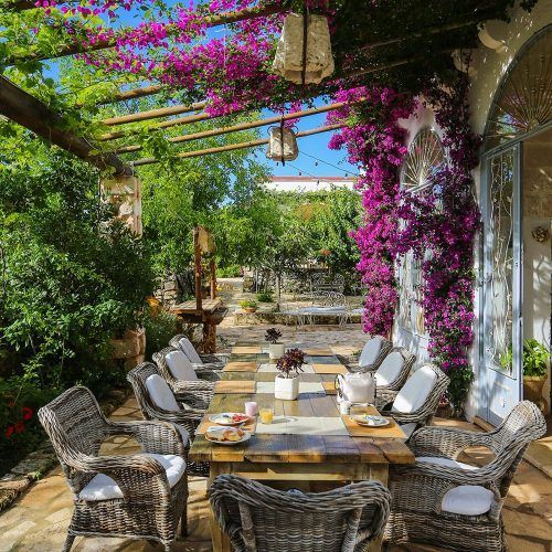 Flower-filled outdoor dining patio with a long table and cushioned chairs under a sunny pergola