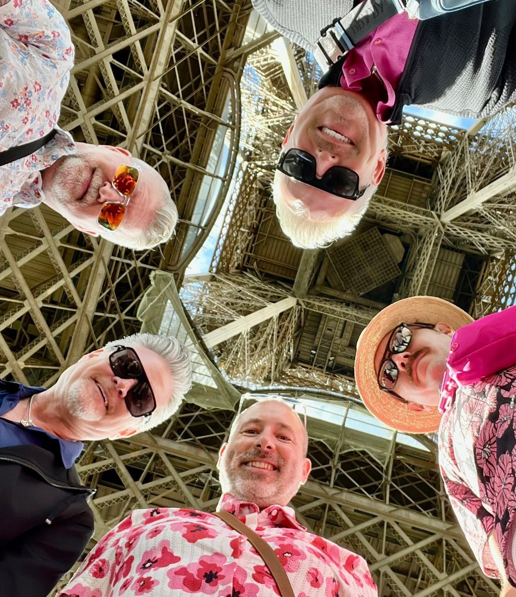 Four people in sunglasses and bright clothing looking down at the camera under a metal lattice ceiling.