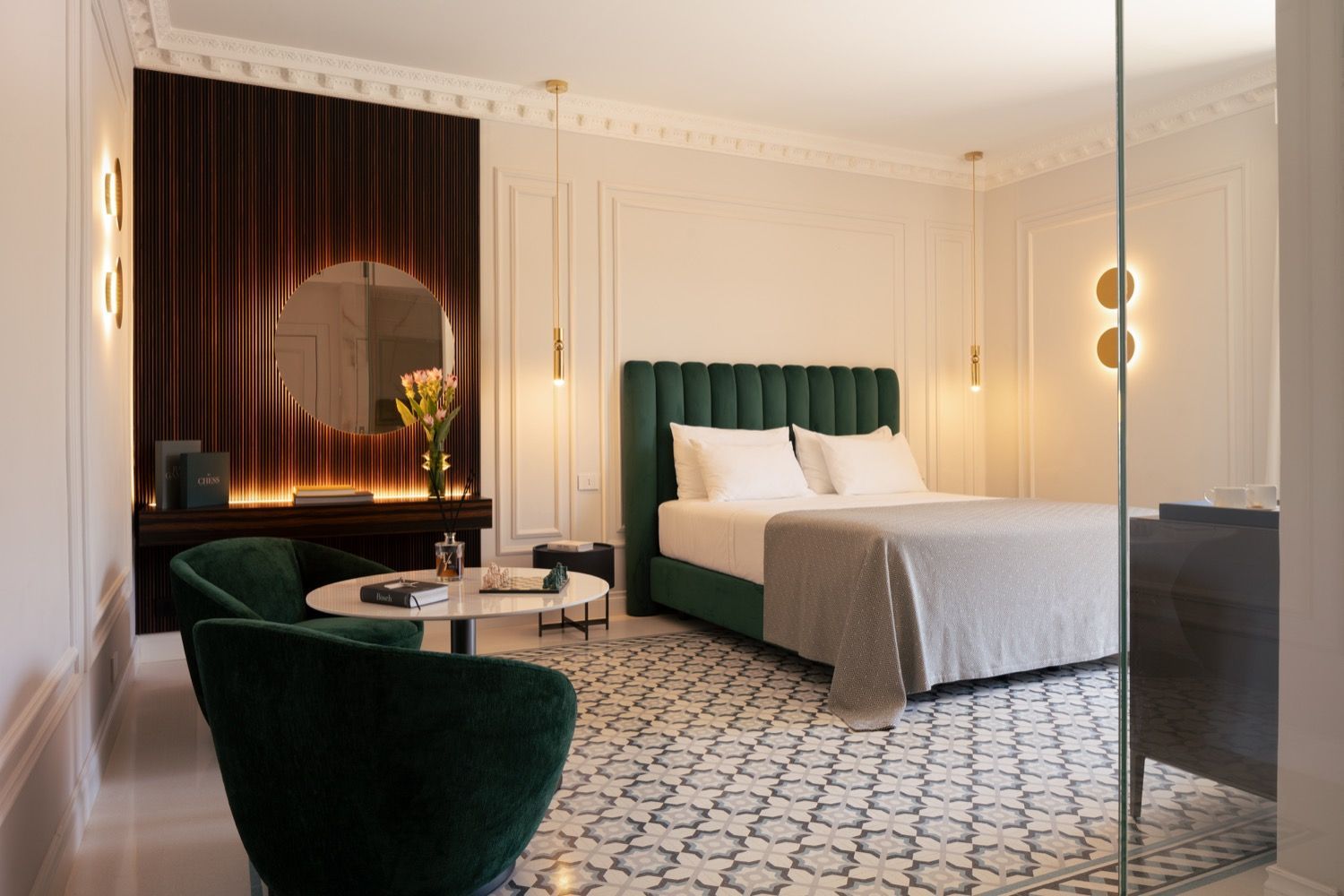 Elegant bedroom with green velvet bed, round mirror, patterned floor, and warm chandelier lighting