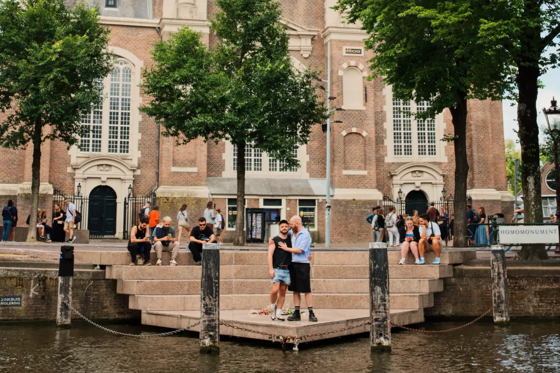 People standing on a canal walkway in front of a historic brick building with trees and water