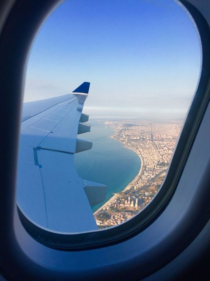 View from airplane window of wing over a coastal city and blue sea under a clear sky