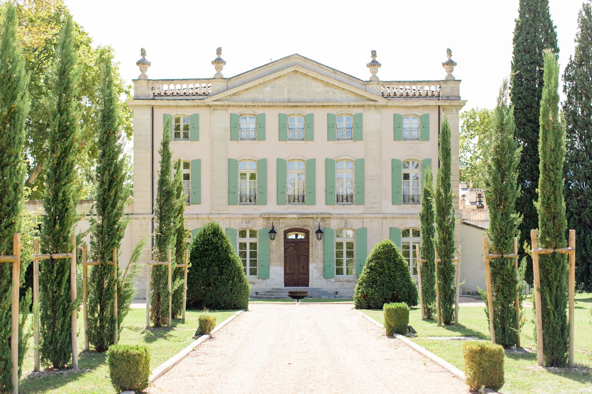 Elegant pale green villa with white trim, framed by tall cypress trees and a gravel driveway