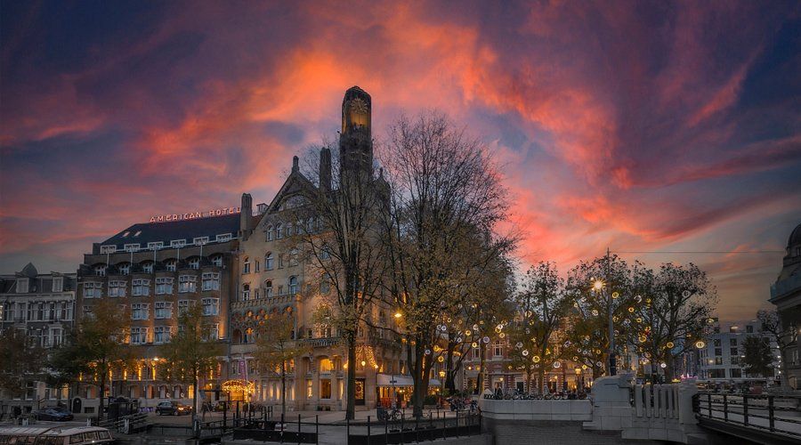 Historic riverside building at dusk with glowing windows beneath a dramatic pink-orange sunset sky