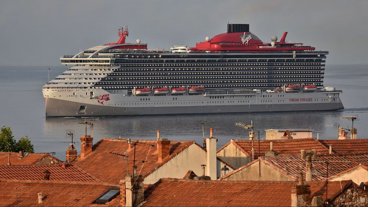 Large white-and-red cruise ship on calm water behind red-tiled rooftops at sunset