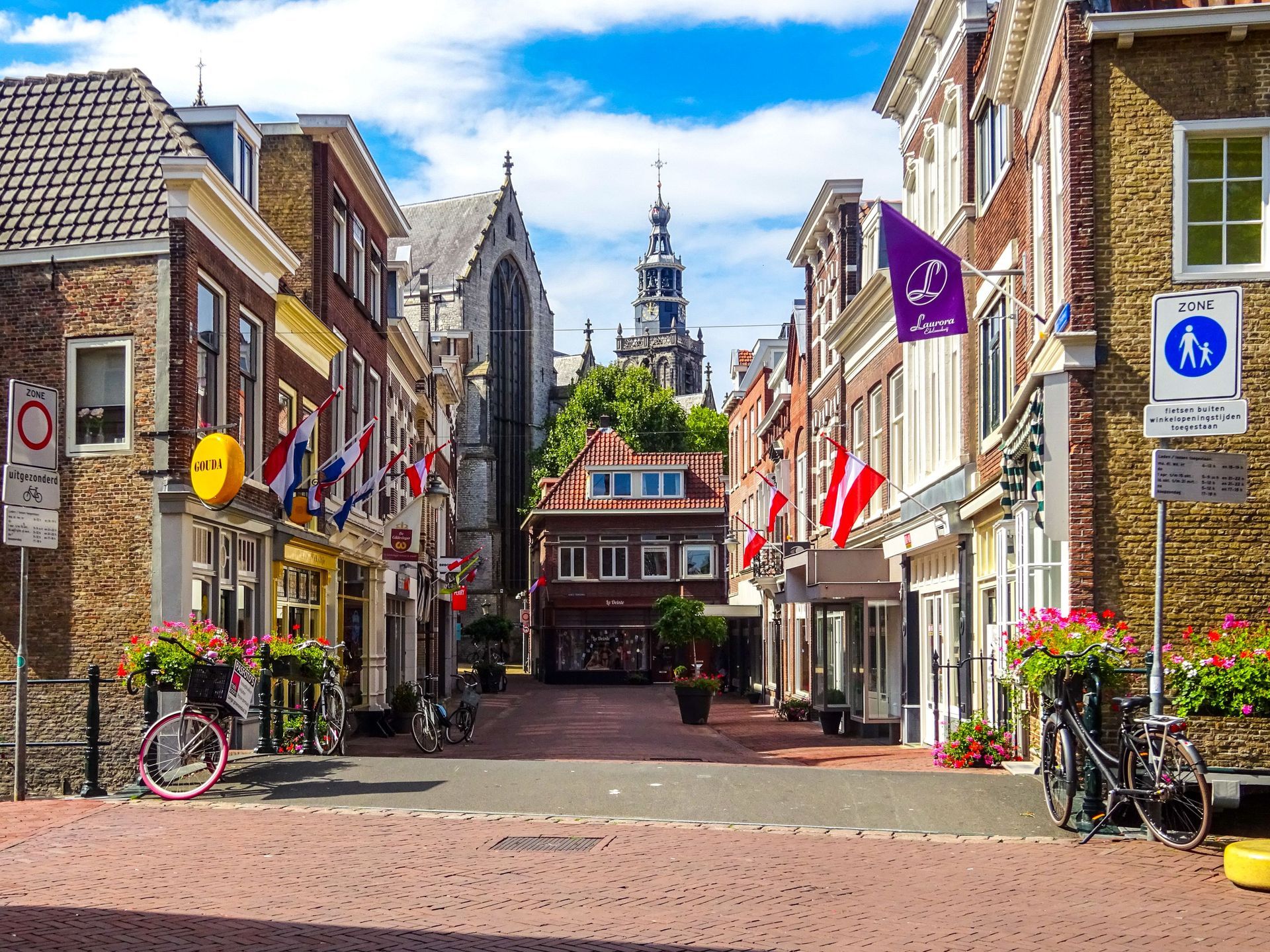 Narrow cobblestone street lined with historic buildings, flags, and bicycles, leading to a church tower under a blue sky