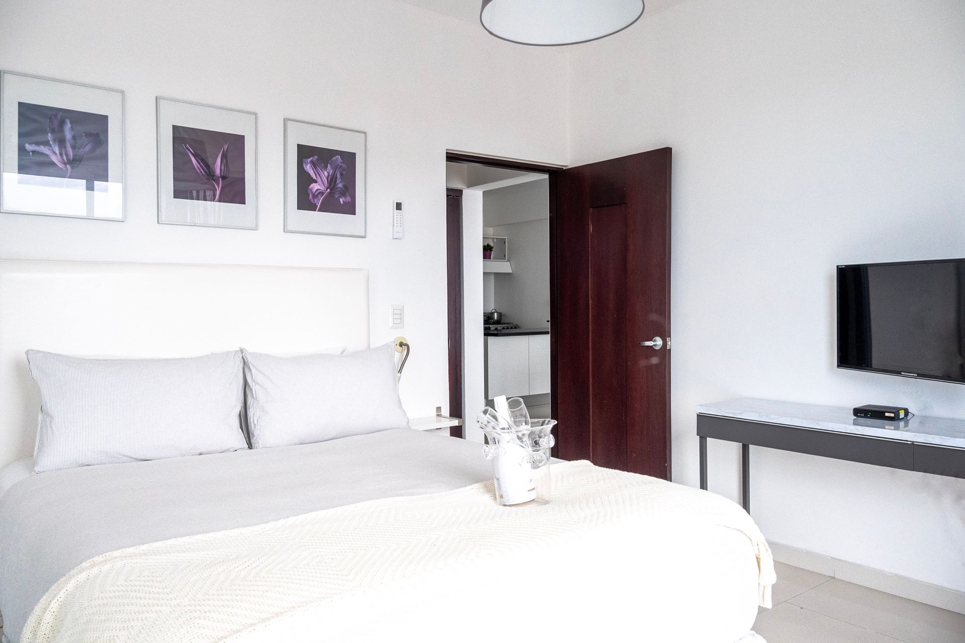 Habitación luminosa de color blanco con cama, cuadro enmarcado, armario abierto y escritorio con televisor.