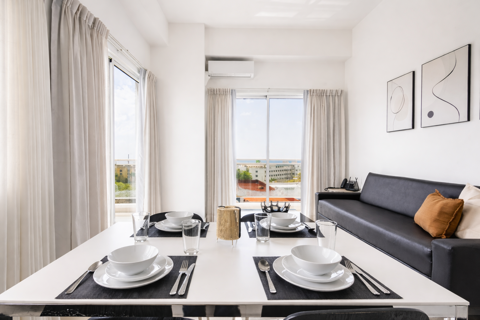 Luminoso salón-comedor con mesa para dos, sofá gris y vistas al balcón a través de grandes ventanales.
