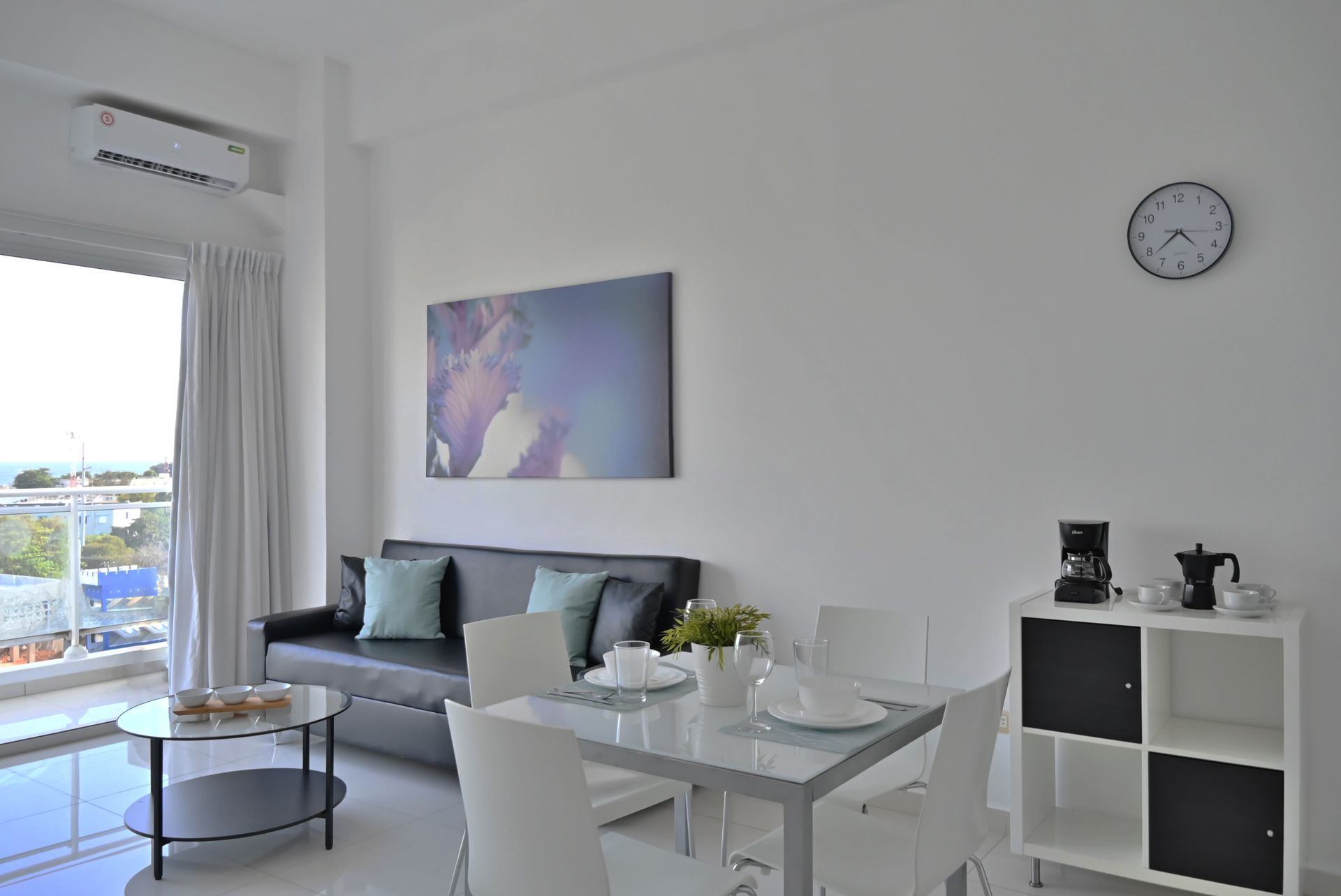 Sala de estar luminosa con sofá gris, mesa de comedor de cristal, cuadros en la pared y una pequeña estantería cerca de una ventana.