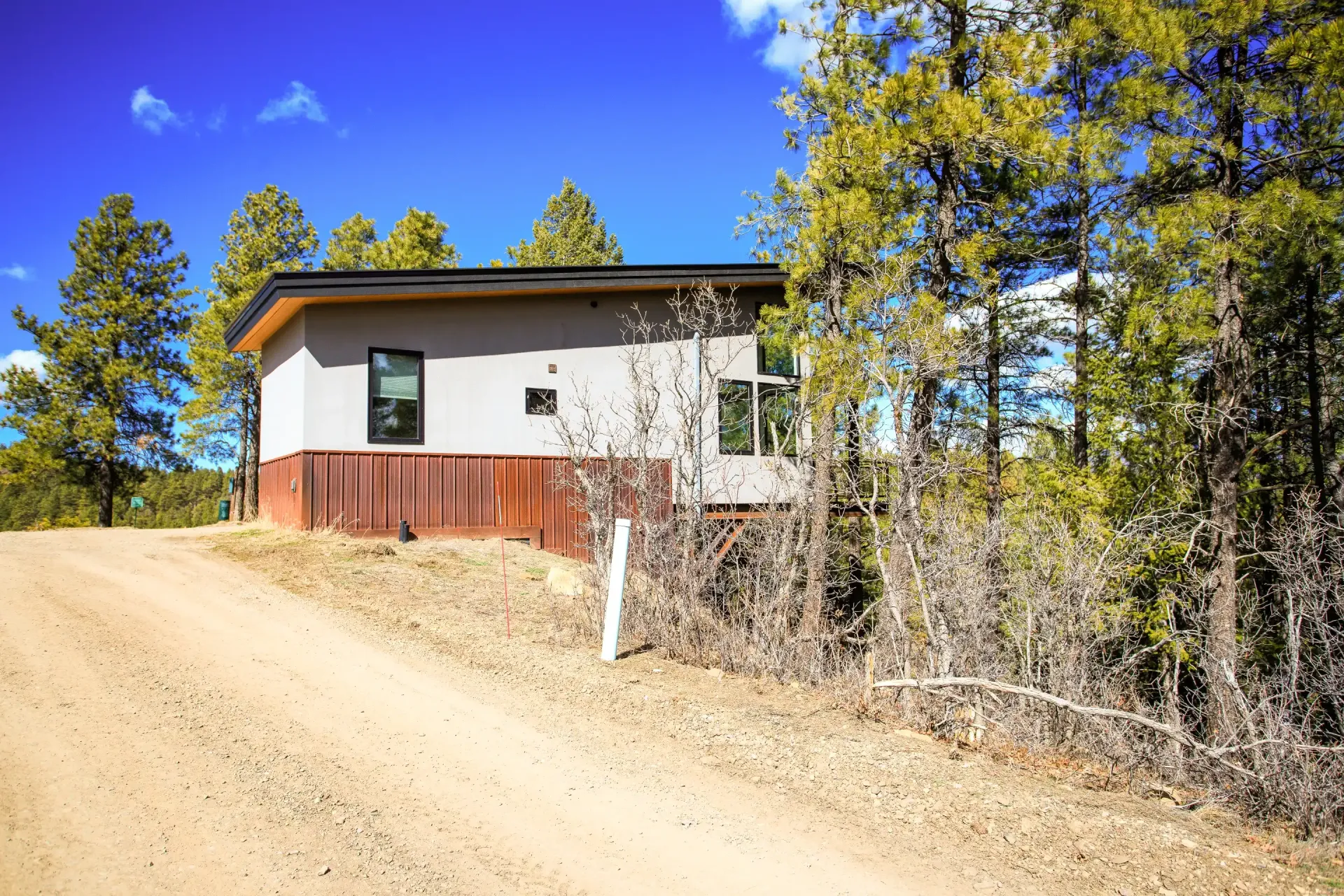 A modern cabin with grey walls and wood siding sits on a dirt road, surrounded by pine trees under a clear blue sky.