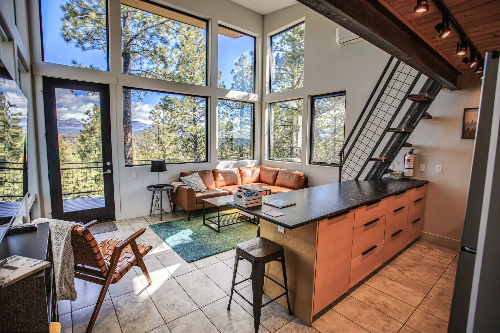 A sunlit, modern living area with floor-to-ceiling windows, a brown leather sectional, a kitchen island, and metal stairs.