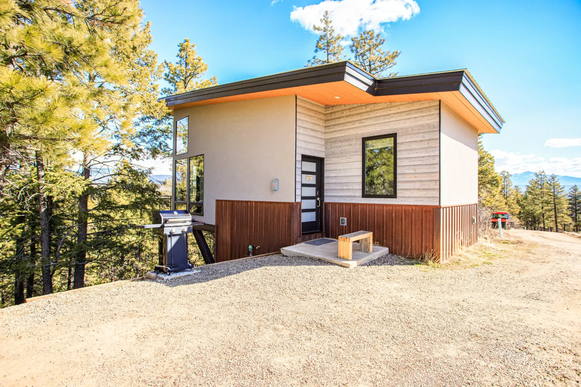 A small, modern cabin with wood and white siding, surrounded by pine trees on a sunny, gravel-covered mountainside.