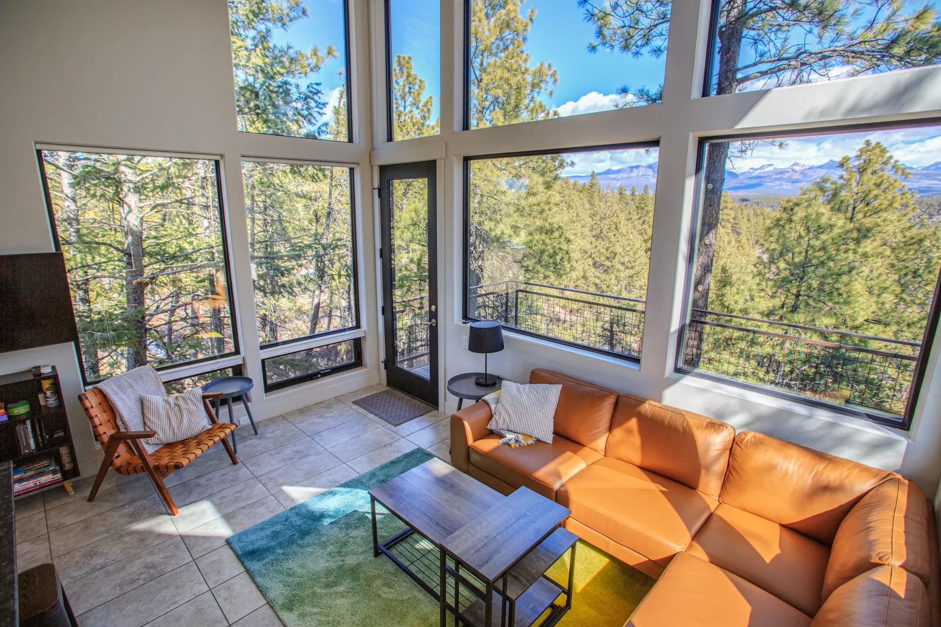 Modern living room with a large tan sectional sofa, accent chair, and floor-to-ceiling windows overlooking a forest.