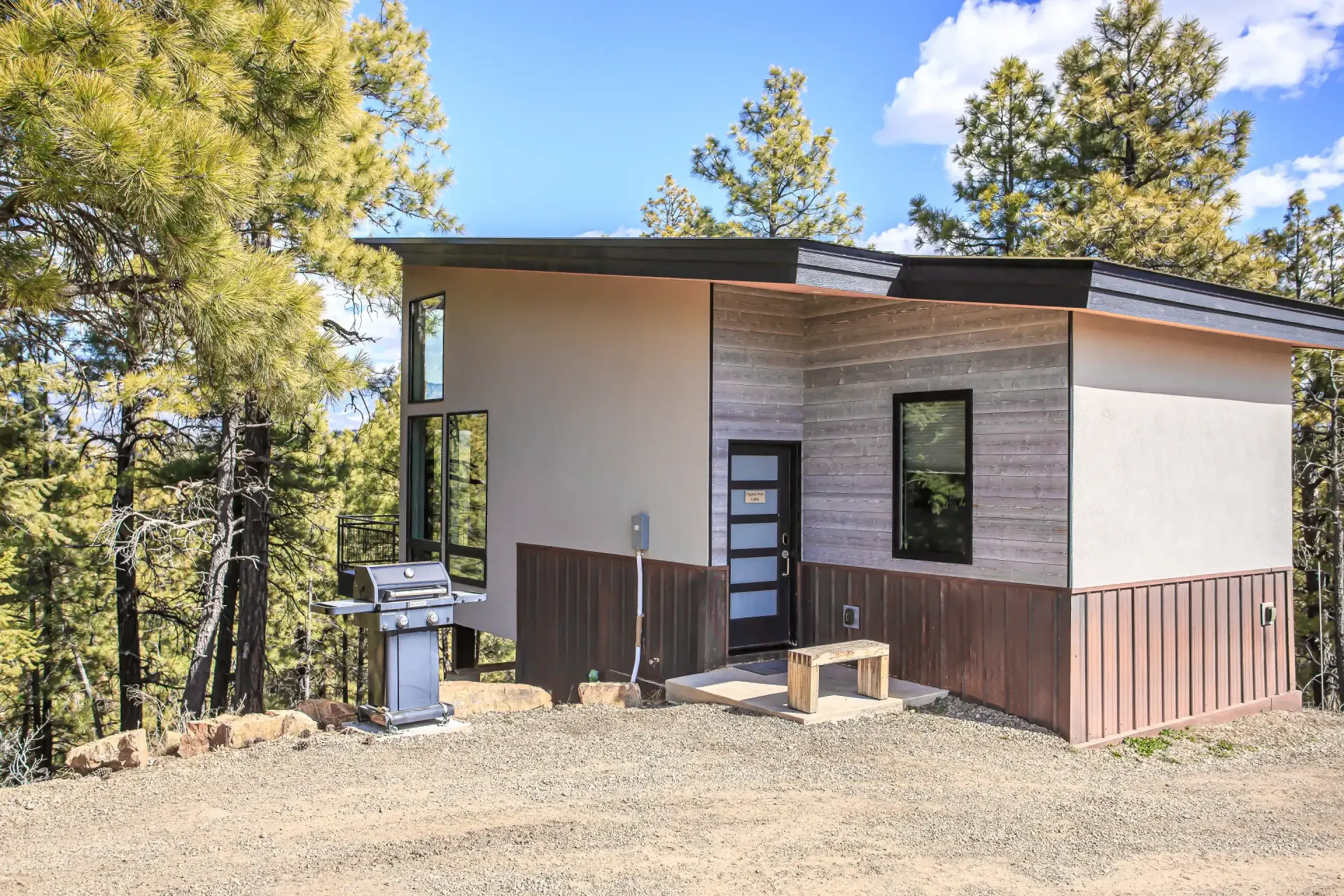 A modern cabin with light beige, wood, and dark vertical siding sits in a pine forest on a gravel lot with a grill.