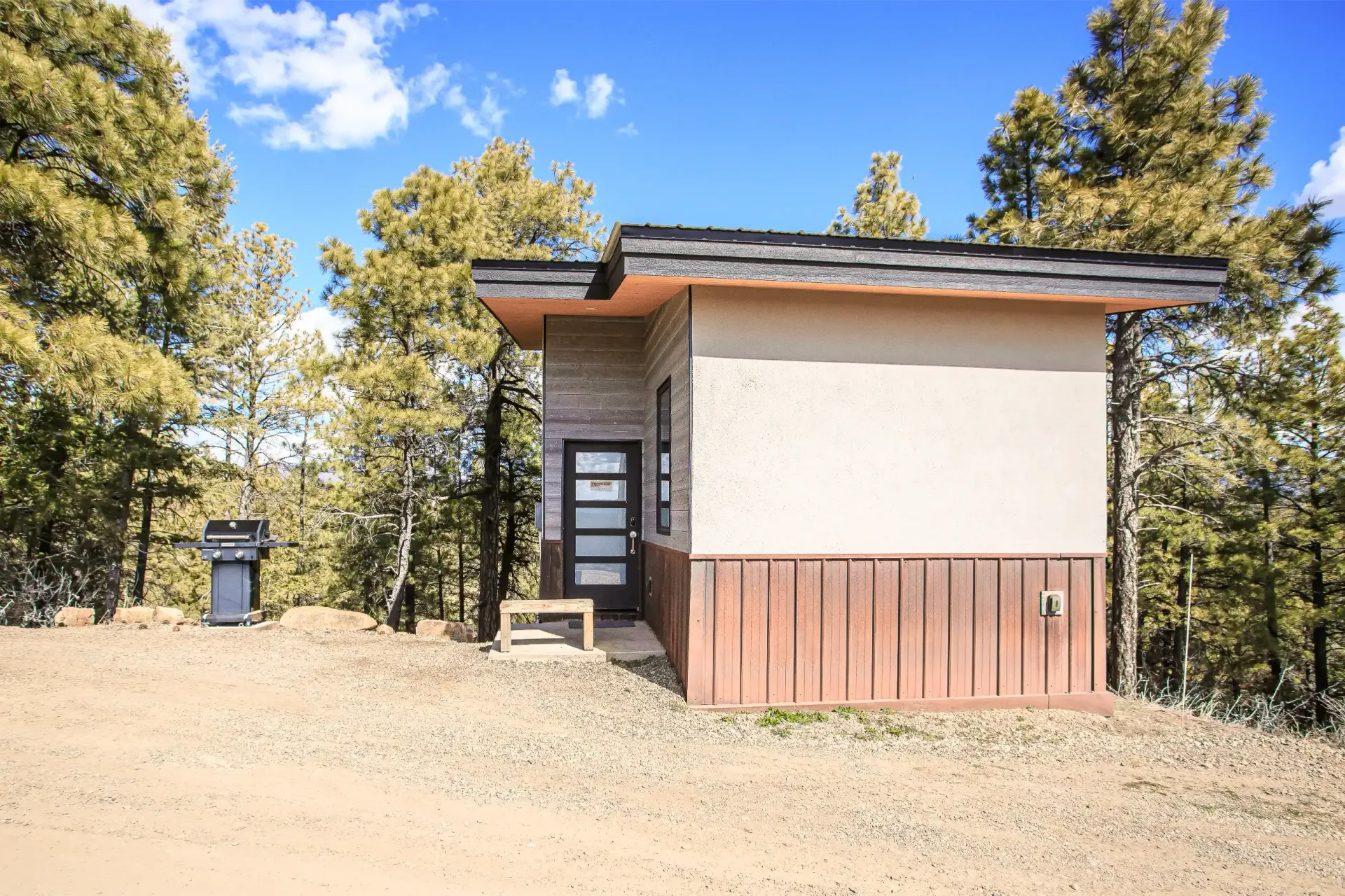 A small, tan cabin with vertical wood siding sits in a forest clearing next to an outdoor grill.