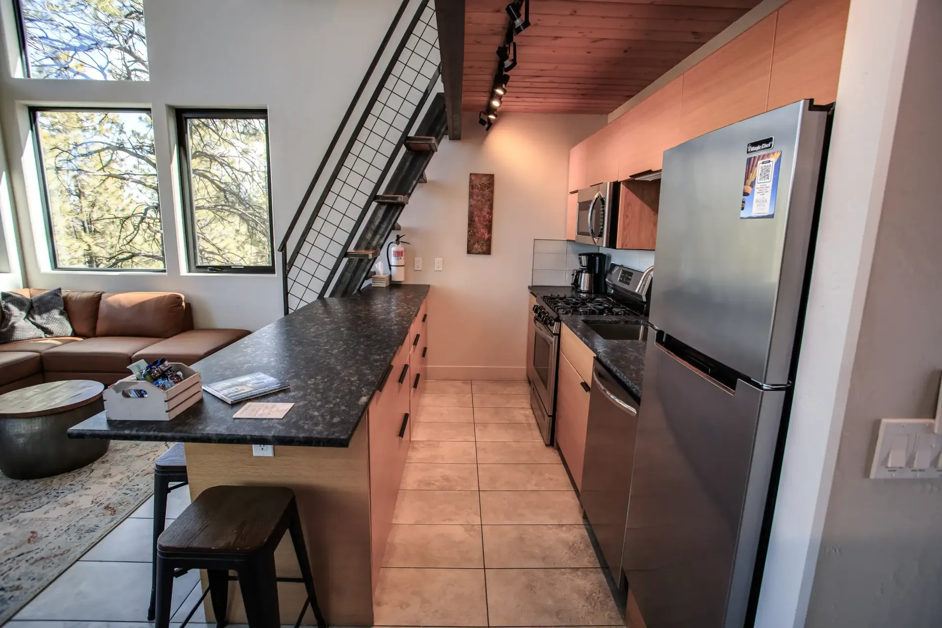 A view of a modern kitchen featuring a dark countertop island, stainless steel appliances, and a staircase.