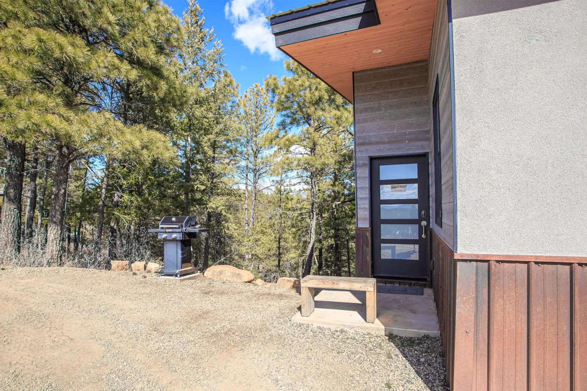 A modern cabin exterior featuring a multi-paned glass door, a small stone bench, and a grill on a gravel patio near trees.