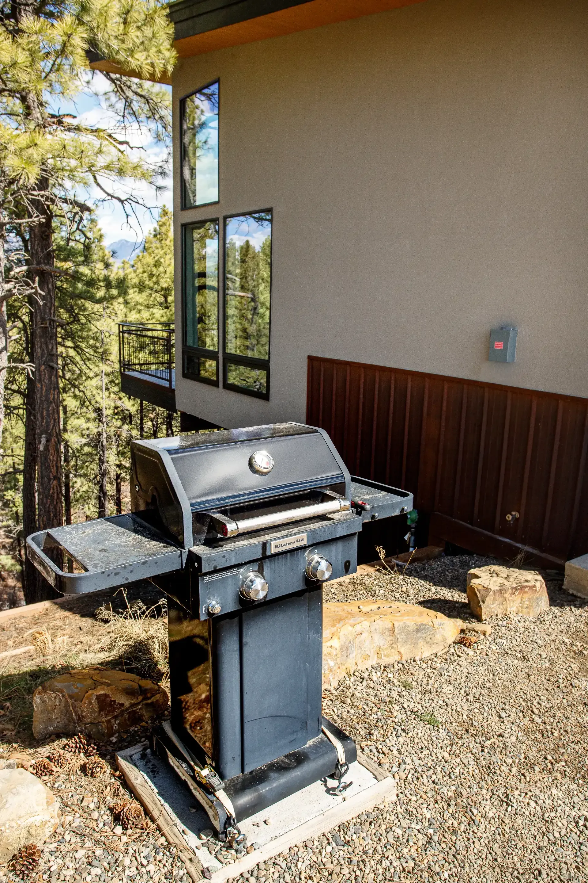 A dark charcoal-colored gas grill stands on a gravel patio outside a house with tan stucco and dark wood paneling.