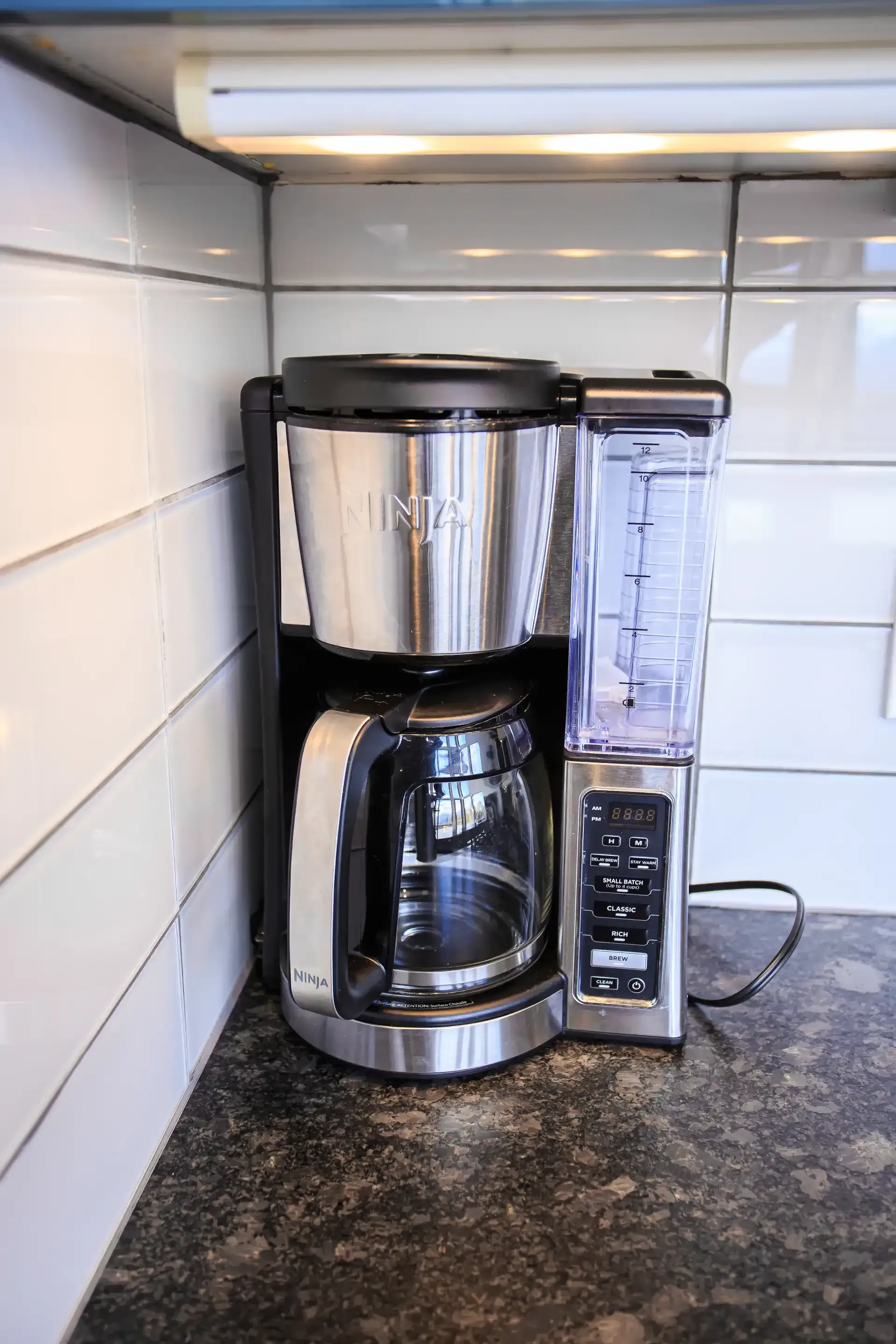 A Ninja coffee maker sits on a dark countertop in a kitchen with white subway tile walls.