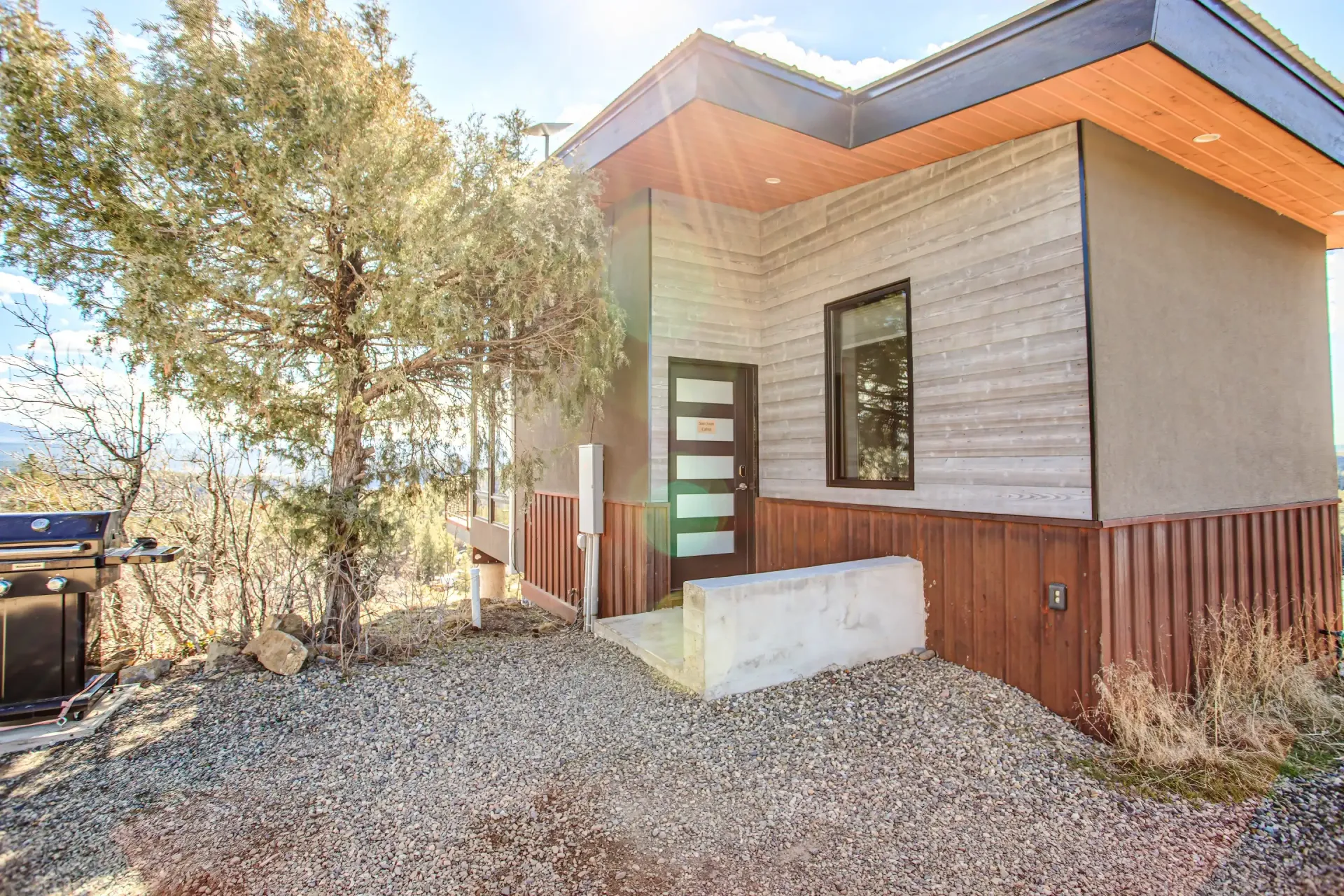 A modern, small cabin with layered siding and a rusted metal base sits on a gravel lot under a bright, sunny sky.
