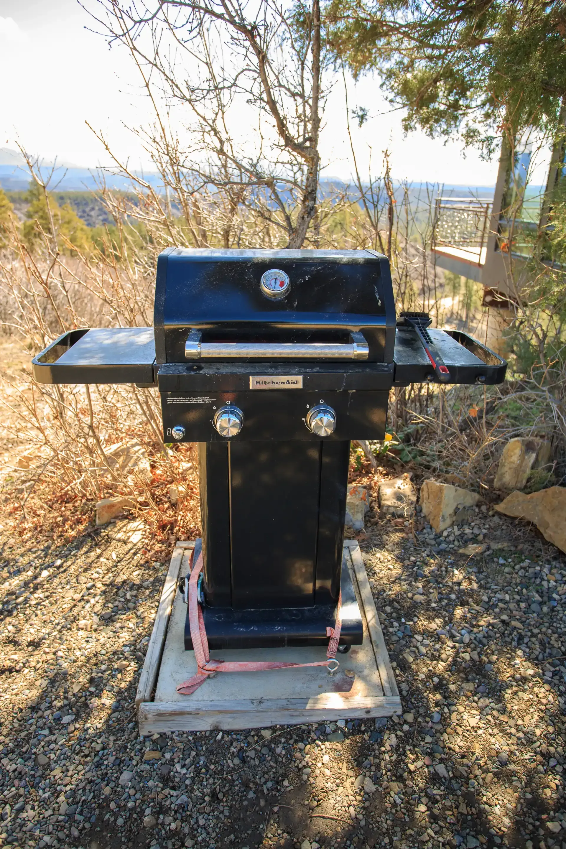 A black two-burner gas grill secured to a wooden base on a gravel surface outdoors against a backdrop of trees and hills.