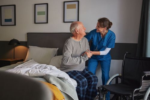 Caregiver in blue helping a seated patient in a bedroom, with bed, lamp, and framed art in the background