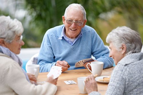 Three seniors smiling and playing cards at a wooden table with coffee cups outdoors