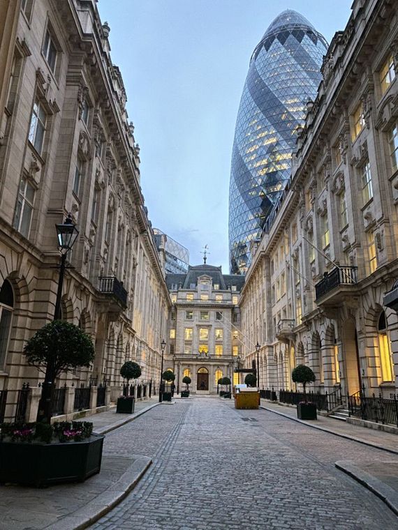 Cobblestone street between historic stone buildings, with the Gherkin tower rising in the background at dusk.