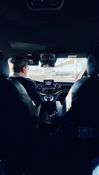 Inside a car, two people sit in front seats, seen from the back with the road ahead through the windshield.