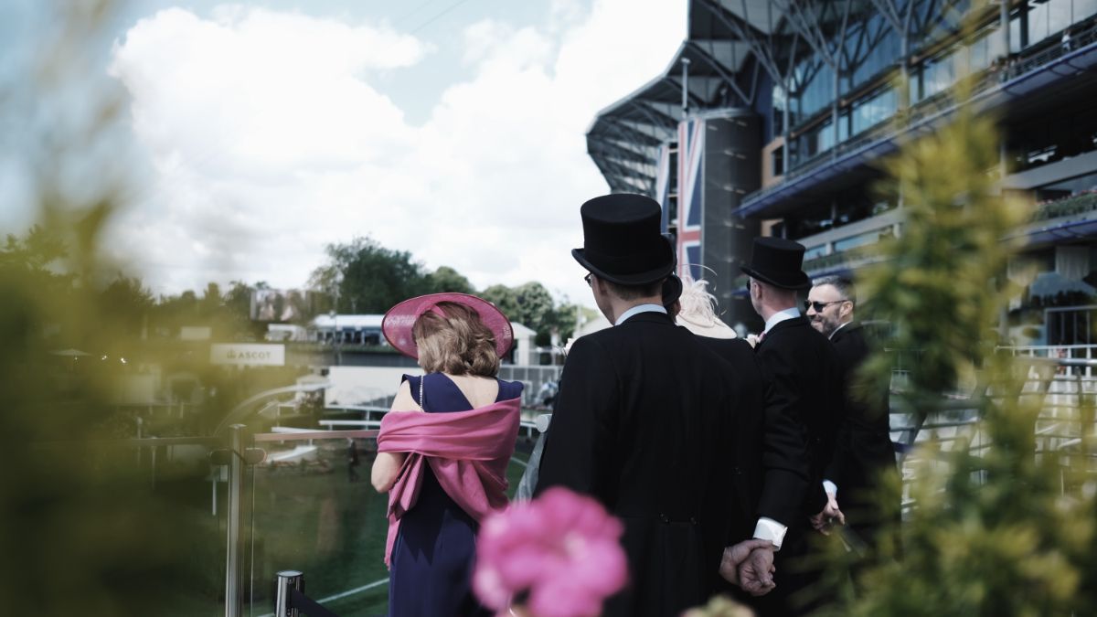 Formal guests in black suits and pink attire walk outdoors near a modern glass building.