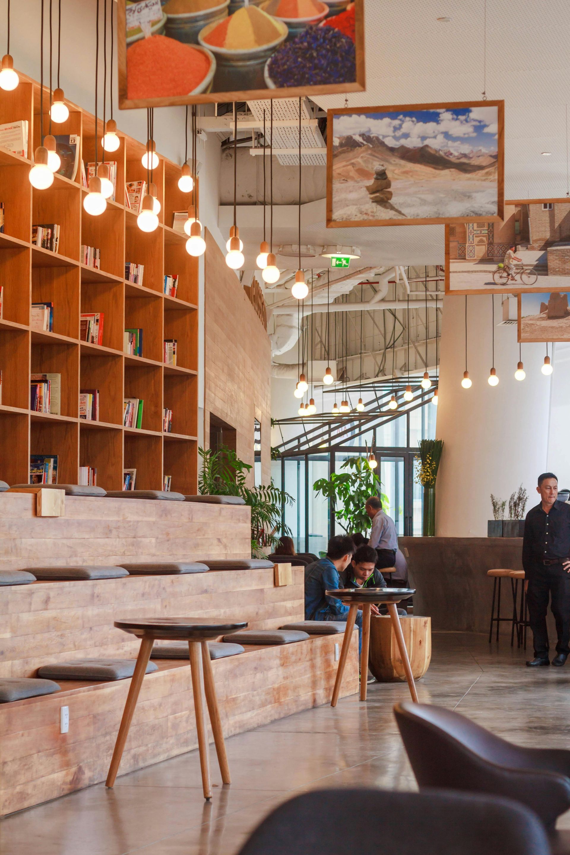 Wooden bookshelves, seating, and tables in a bright interior with artwork, people, and hanging lights.