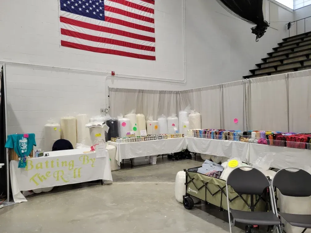 Booth tables with items for sale at an indoor fair beneath a large American flag