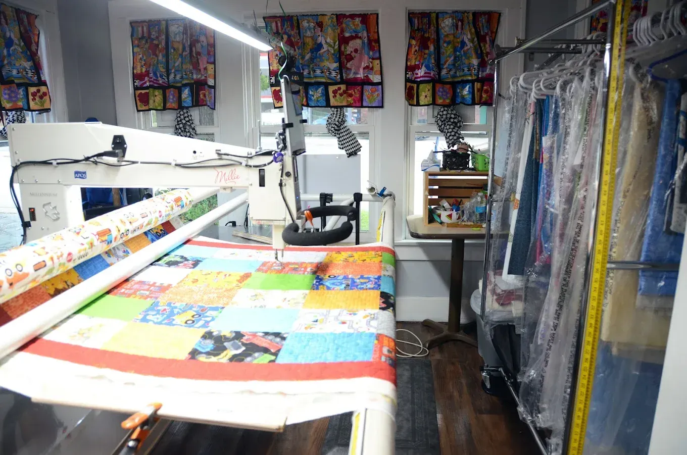 Quilting studio with a sewing machine, colorful quilt on a frame, and hanging fabric rolls