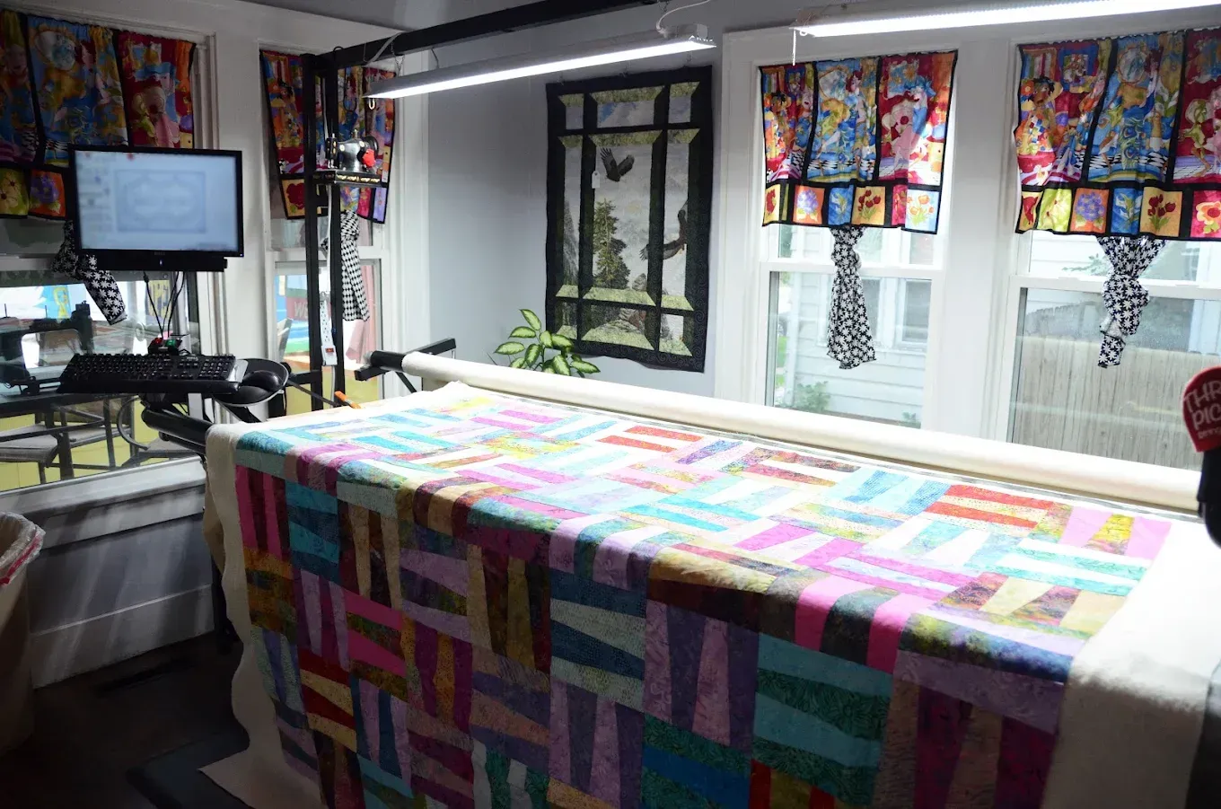 Colorful quilt displayed on a table in a sewing room with windows and hanging fabric samples.