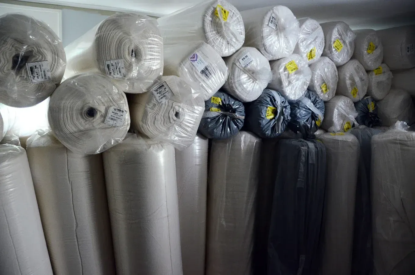 Stacks of rolled plastic-wrapped materials in white, black, and gray, stored in a warehouse room.