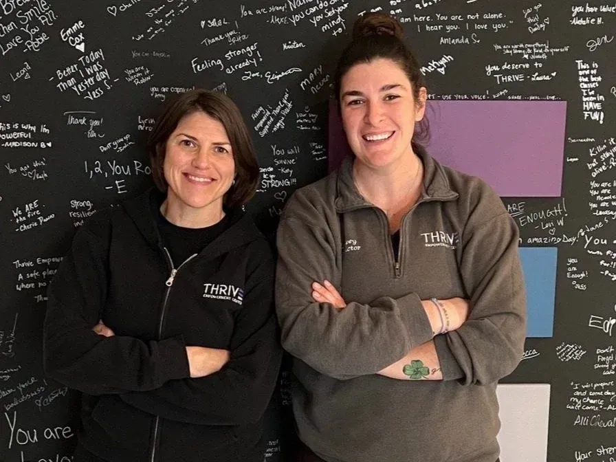 Two staff members standing with arms crossed in front of a chalkboard wall, smiling