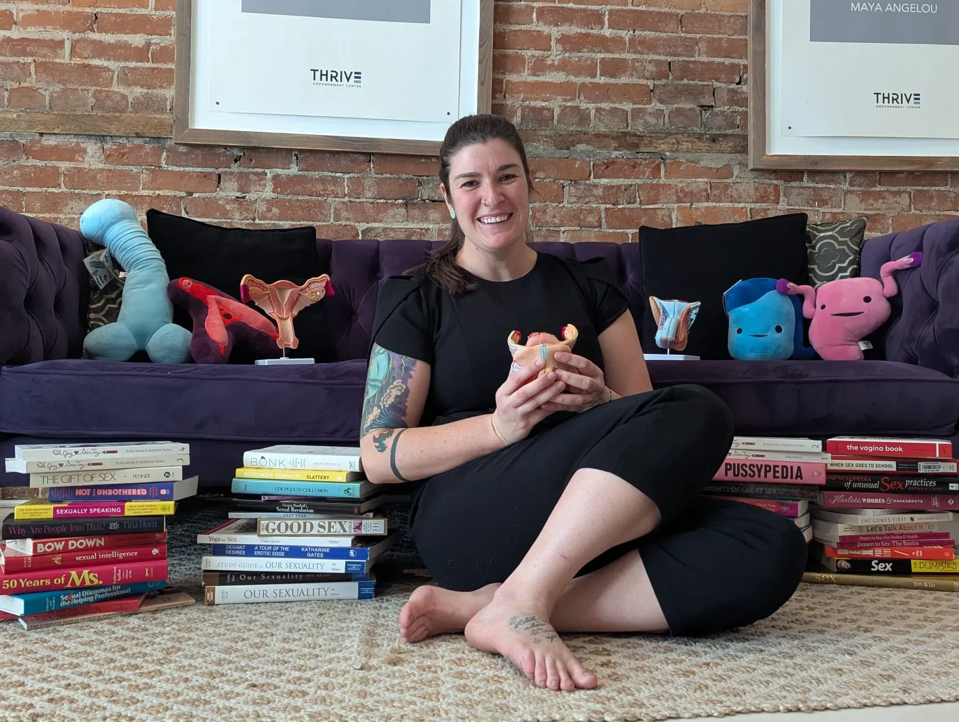 Person sitting cross-legged on a rug in front of a couch, smiling, with colorful plush toys and stacked books.