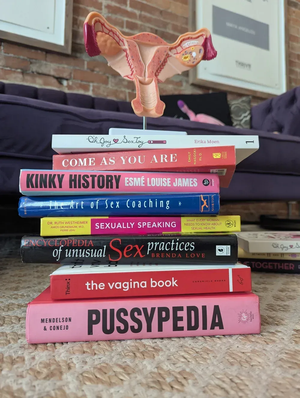 Stack of colorful sex-positive books with a uterus model on top, on a carpeted floor